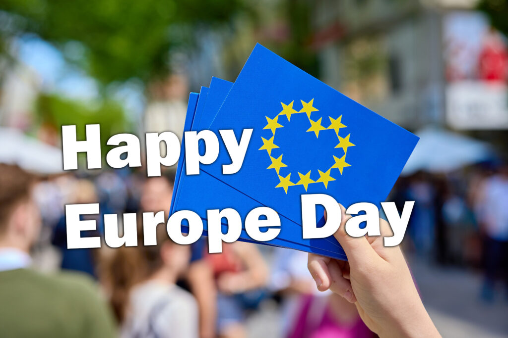 Im Hintergrund hält eine Hand eine Postkarte in der Hand auf der auf blauem Hintergrund ein Herz aus Europasternen zu sehen ist, im Stil der Europaflagge. Im Vordergrund ist der Text "Happy Europe Day"