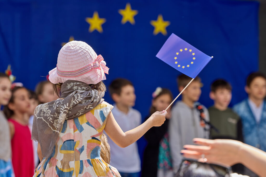ein Kind von hinten auf den Schultern einer nicht sichtbaren Person sitzend. In der Hand hält das Kind eine Europaflagge