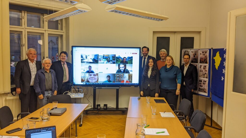 8 Personen neben einem Fernseher, mit Bildern der Teilnehmer in der Zoom-Konferenz