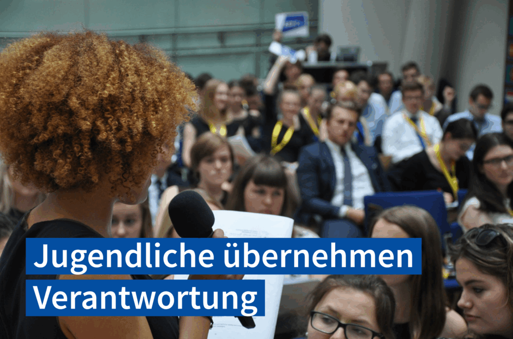 Farbfoto: Im Hintergrund sieht man Menschen sitzend, die einem Vortrag folgen. Im Vordergrund steht eine junge Frau mit einem Mikrophon in der Hand und sie spricht gerade. Text im untern Bildbereich: Jugendliche übernehmen Verantwortung