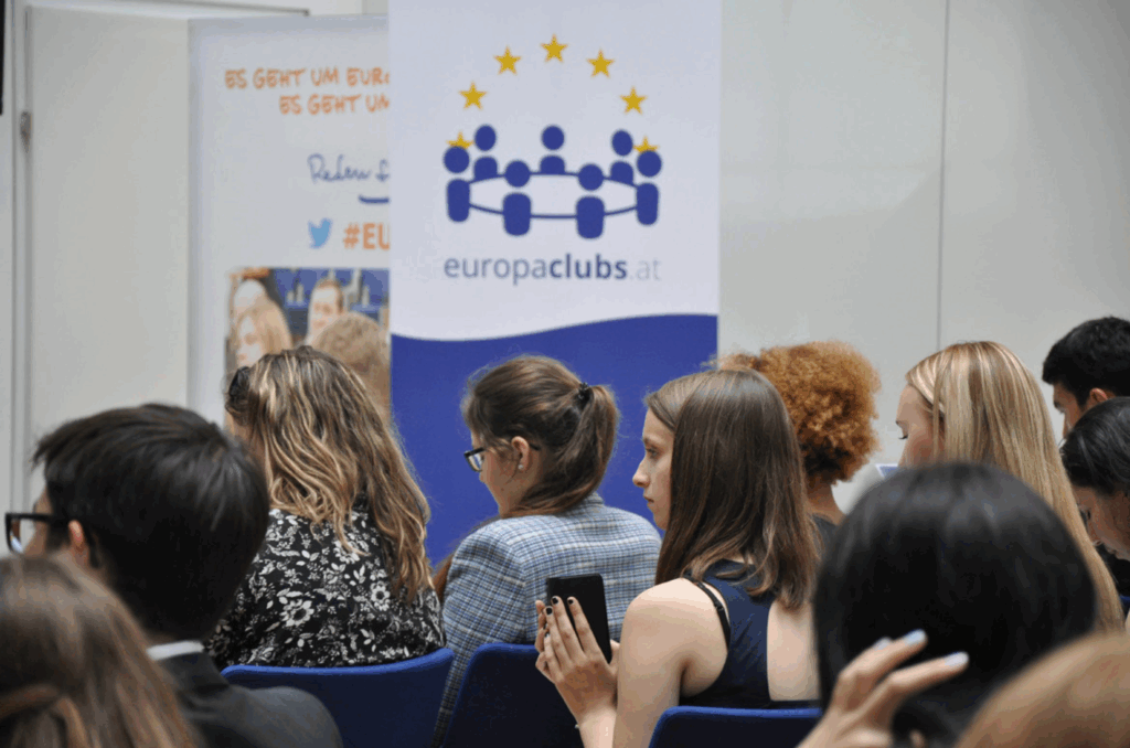 Farbfoto: junge Menschen sitzen auf Sesseln und verfolgen einen Vortrag. Im Hintergrund steht ein Rollup mit dem Logo der "Europaclubs"